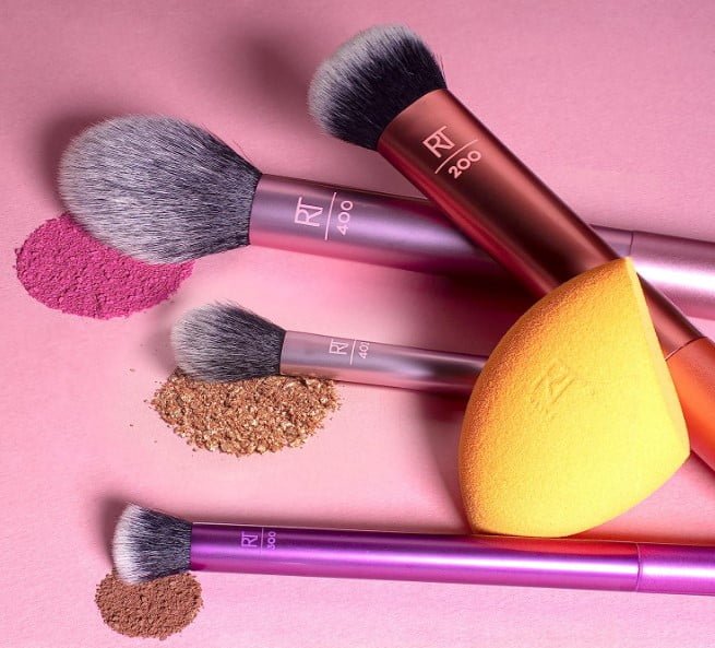 Cuatro brochas y una esponja con polvo de REAL TECHNIQUES Kit Completo De Brochas sobre fondo rosa